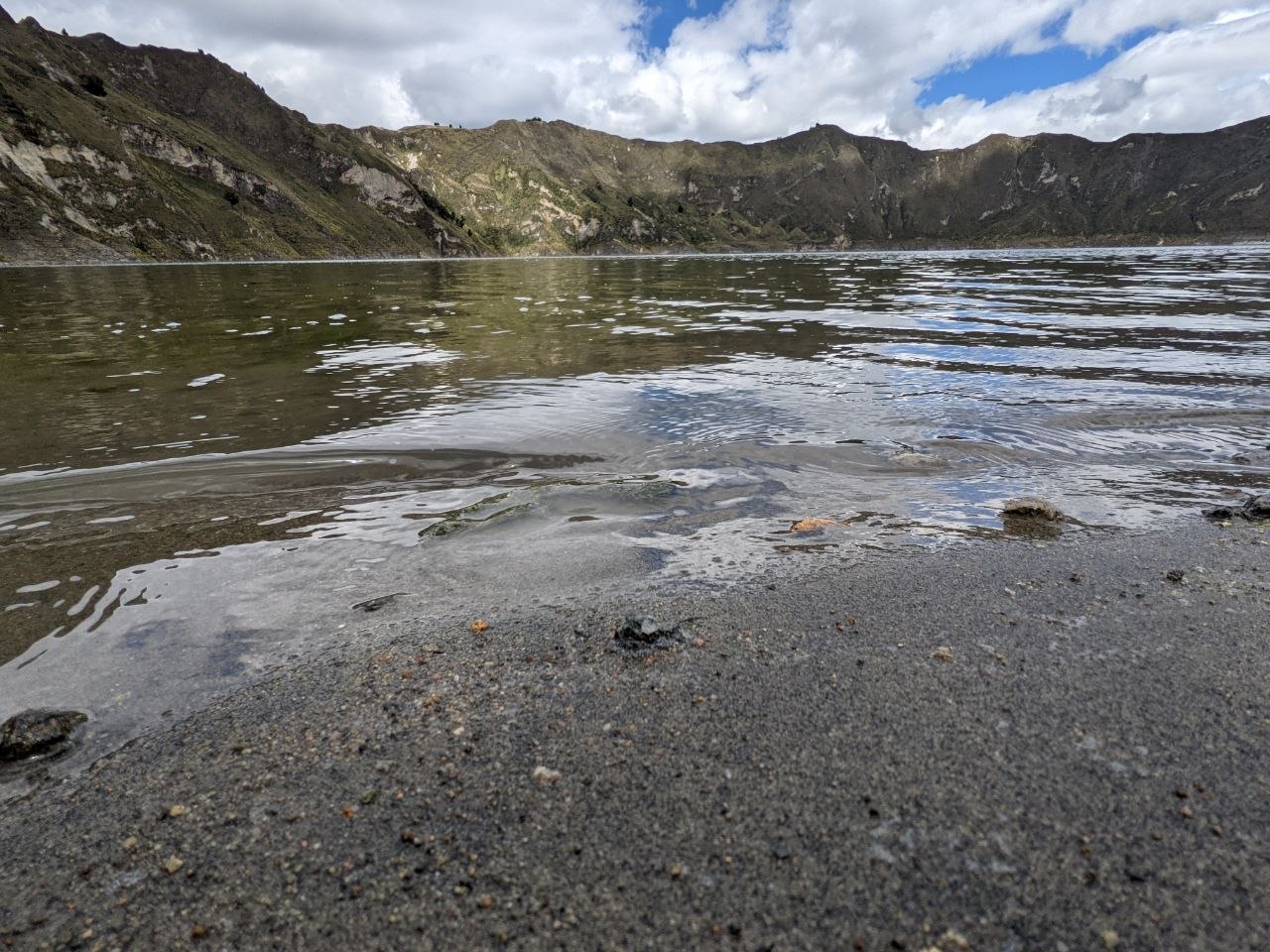 Black volcanic sand shoreline of Laguna Quilotoa with crater walls rising behind the turquoise lake