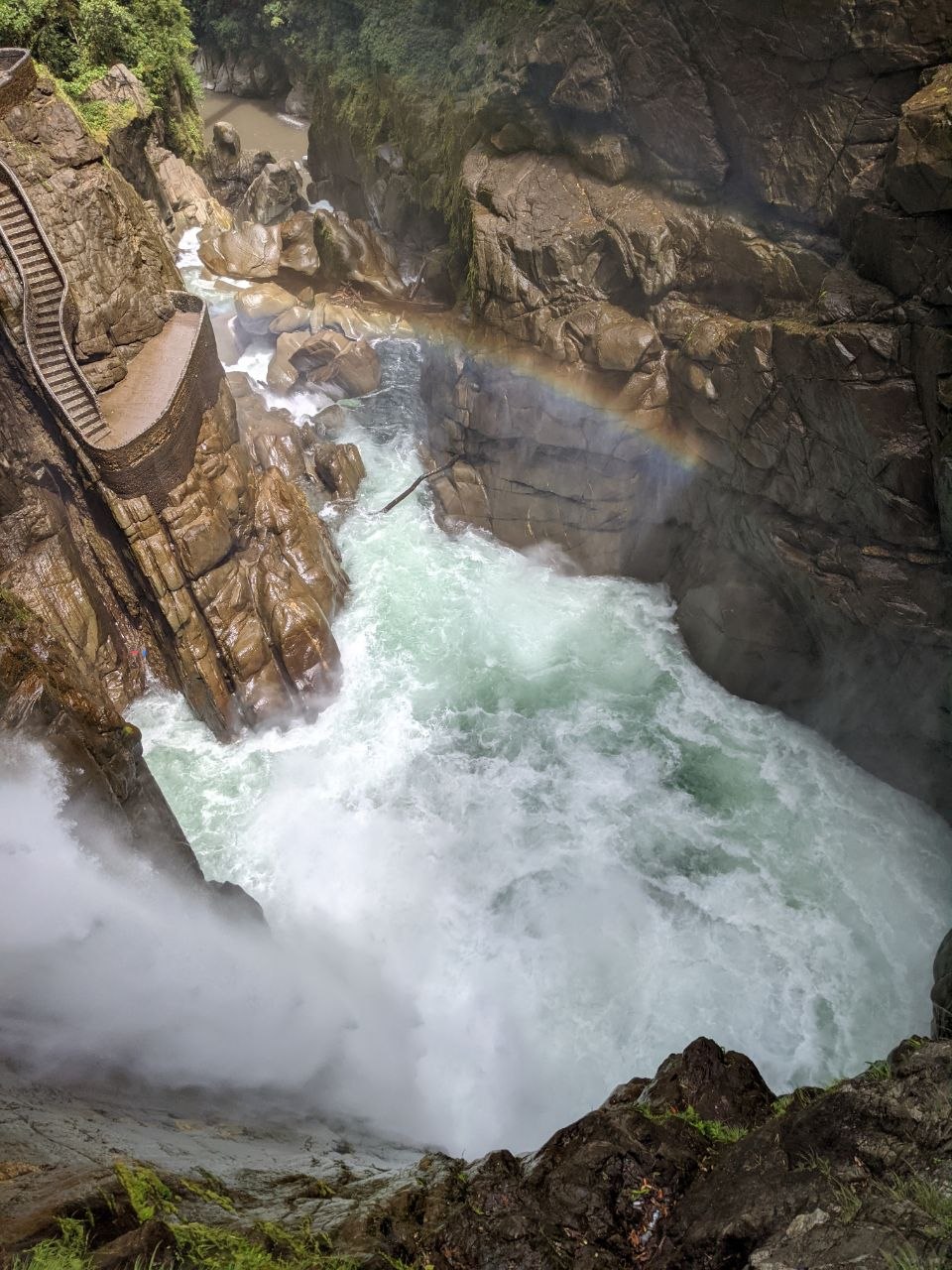 Looking down into the churning pool at the base of Pailón del Diablo with a rainbow visible in the spray and staircase on the left