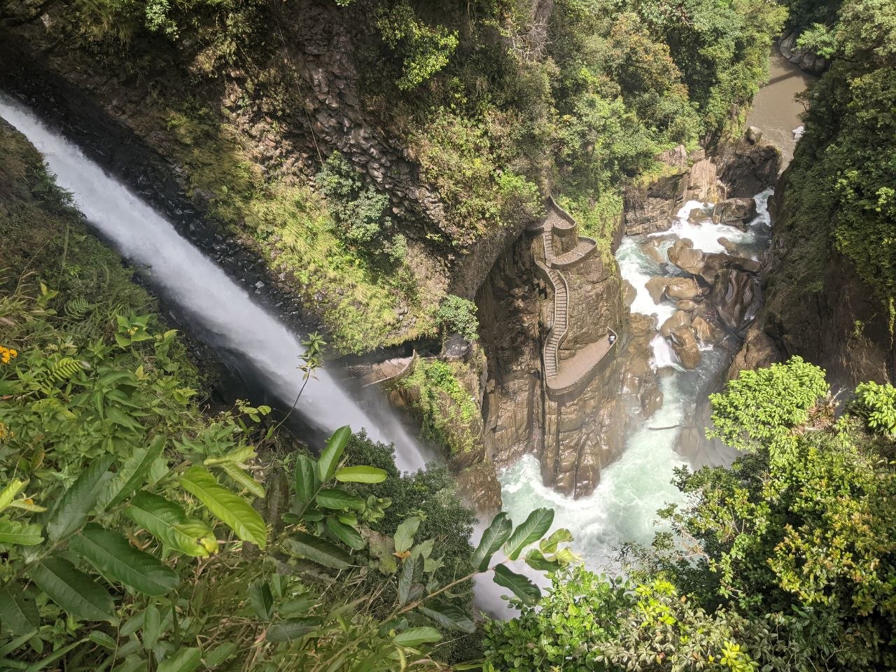 Side angle view of Pailón del Diablo from above showing the waterfall, spiral staircase platform, and raging river below surrounded by tropical vegetation