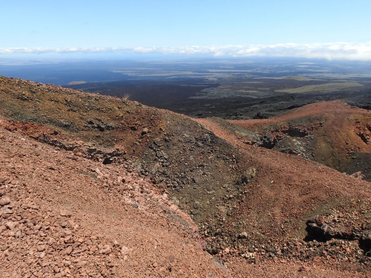 Barren volcanic ridge on the Galápagos Islands with red and black lava terrain dropping to an expansive lava plain and the Pacific Ocean in the distance