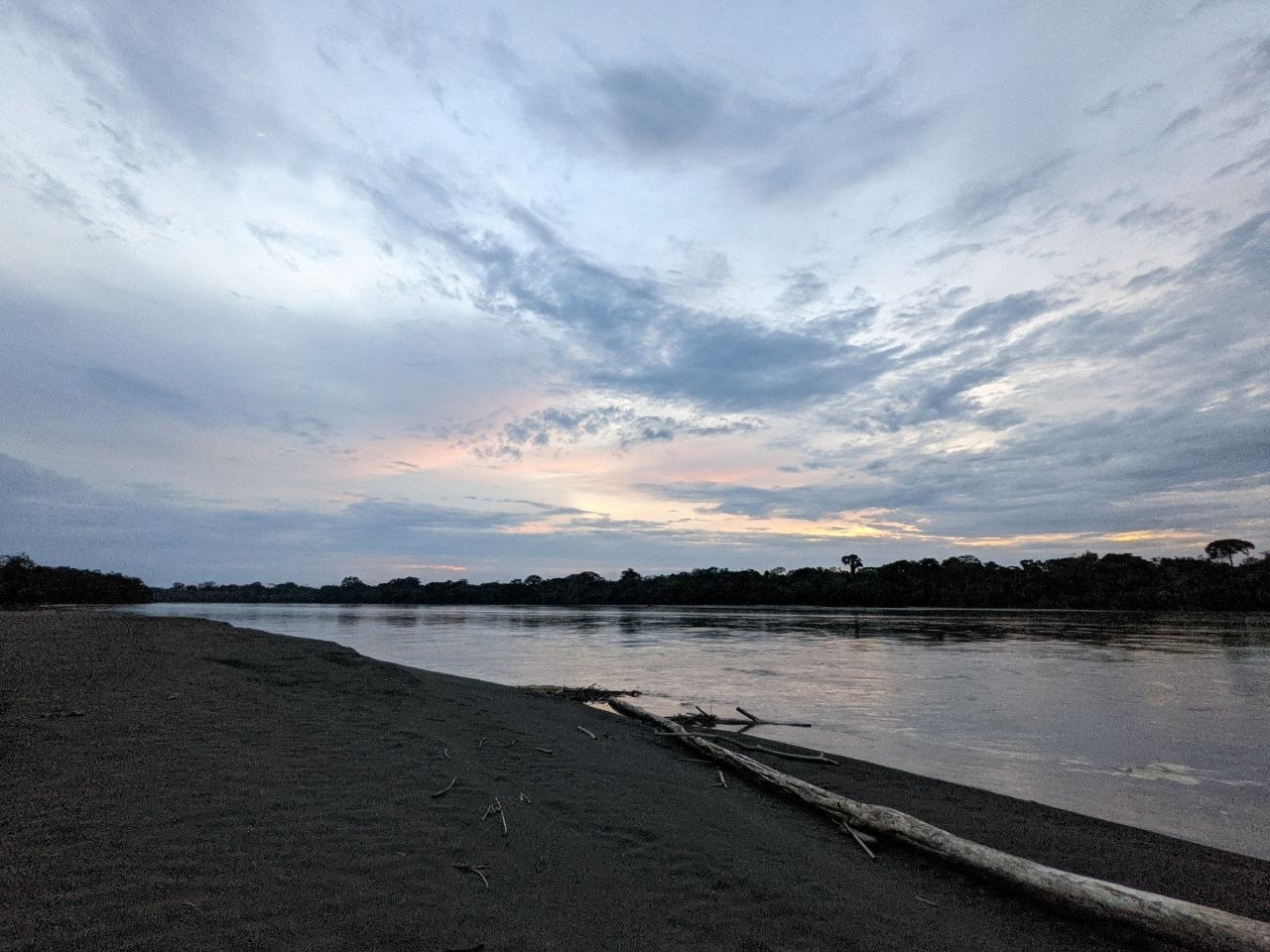 A wide river at dusk in Cuyabeno Wildlife Reserve, with a dark sandy bank in the foreground, driftwood, and the dense jungle silhouetted against a pastel sky
