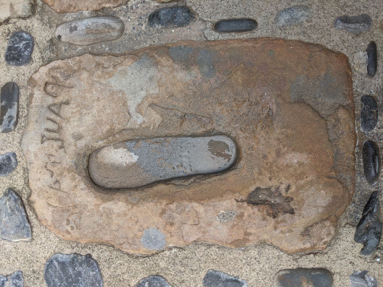 Close-up of a worn stone slab embedded in the cobbled path at San Juan de Gaztelugatxe, with the name carved into its surface and a smooth black pebble set into its centre