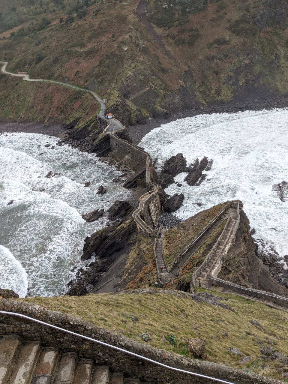 Aerial view looking down from the hermitage at San Juan de Gaztelugatxe showing the narrow stone causeway and switchback staircase threading between crashing waves and jagged black rocks