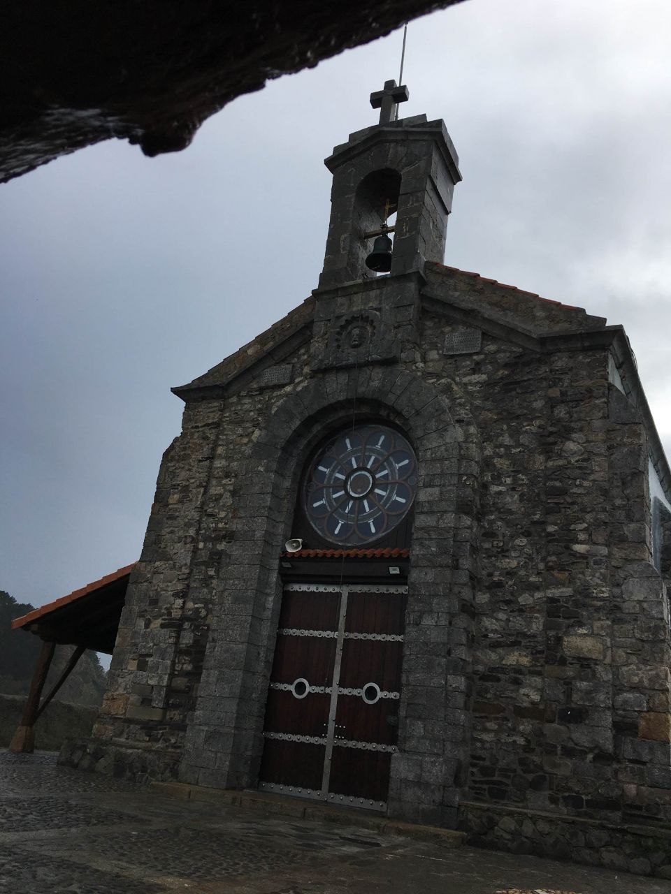 The stone facade of the Hermitage of San Juan de Gaztelugatxe seen from below, with its wooden door, circular rose window, bell tower, and cross against an overcast sky