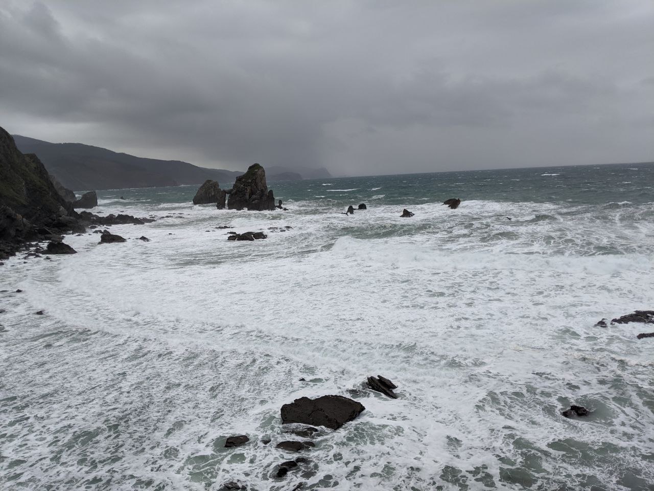 Wild stormy sea at the base of the Gaztelugatxe cliffs with heavy white surf breaking over black flysch rock formations and a jagged sea stack rising from the water under a grey sky
