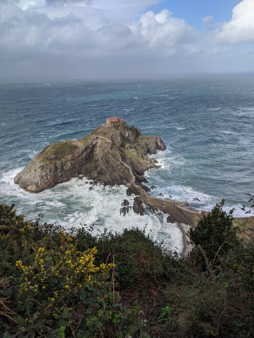 Wide view of San Juan de Gaztelugatxe islet from the clifftop showing the hermitage perched on top, the stone staircase zigzagging up from the causeway, and the stormy Bay of Biscay stretching to the horizon