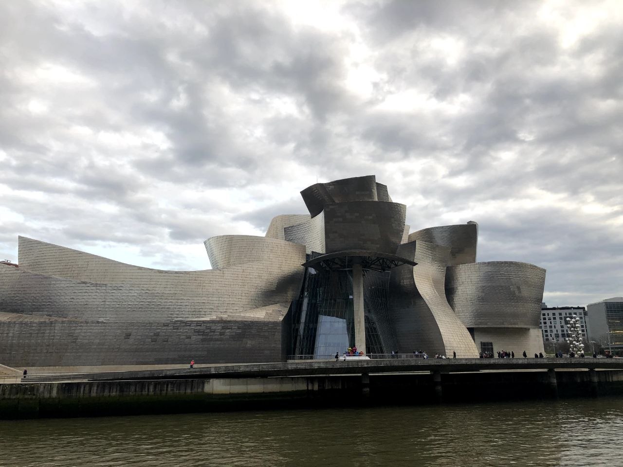 The Guggenheim Museum Bilbao seen from across the Nervión river, its titanium-clad curved forms rising against a dramatic cloudy sky