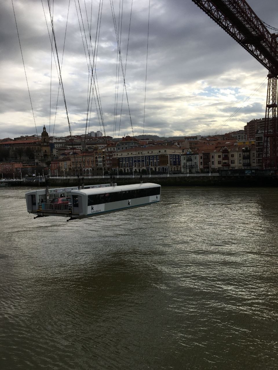 The suspended gondola of the Vizcaya Bridge crossing the Nervión estuary near Bilbao, hanging from steel cables with the town of Portugalete and its colourful buildings in the background