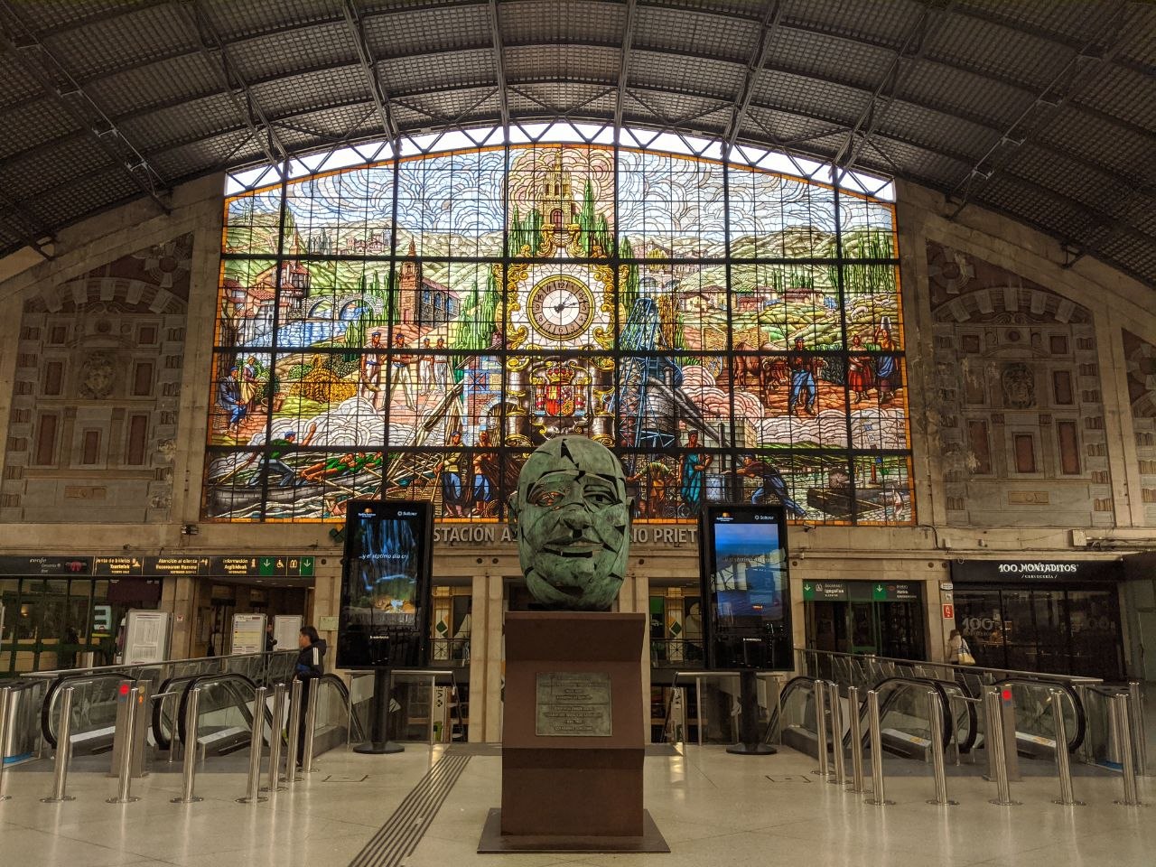 The interior of Bilbao's Estación de Abando showing the vast stained glass window depicting Basque rural and industrial life above the ticket barriers, with a large bronze sculptural head on a plinth in the foreground
