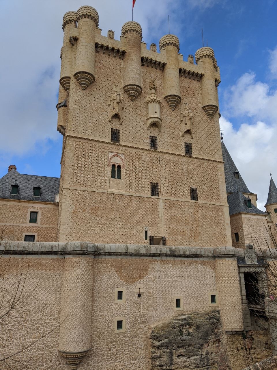 The Torre de Juan II of the Alcázar of Segovia seen from below, its diamond-point stone exterior rising above the outer walls with slate-capped turrets against a blue sky