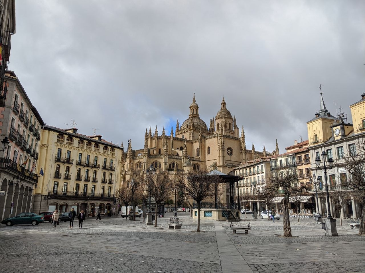 Segovia's Plaza Mayor under an overcast winter sky, with the Gothic Cathedral's pinnacles and dome rising above the surrounding arcaded buildings and bare trees
