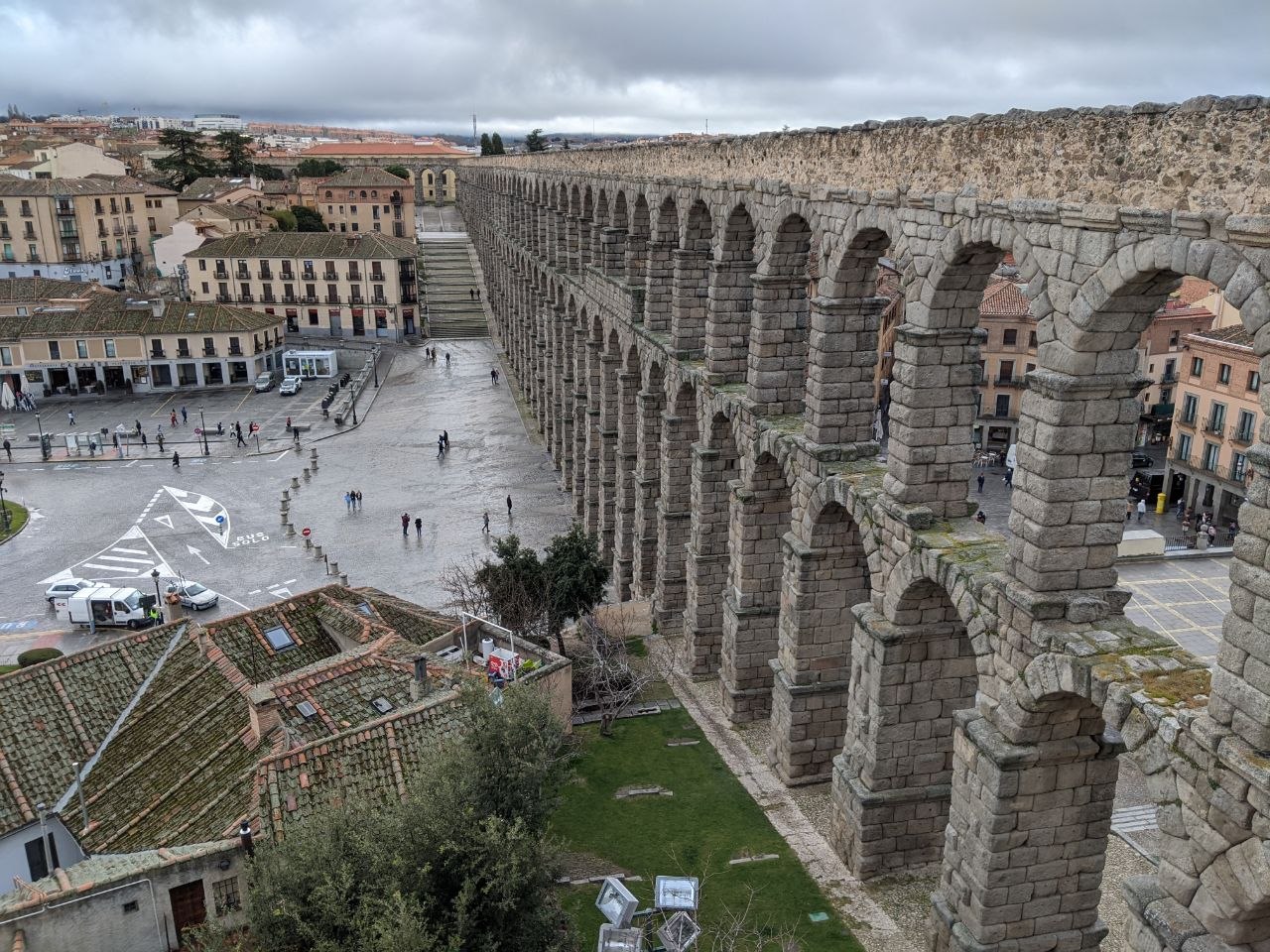 Aerial view along the top of the Roman Aqueduct of Segovia from above, showing the double tier of granite arches stretching across the Plaza del Azoguejo with the old town rooftops below under a grey sky