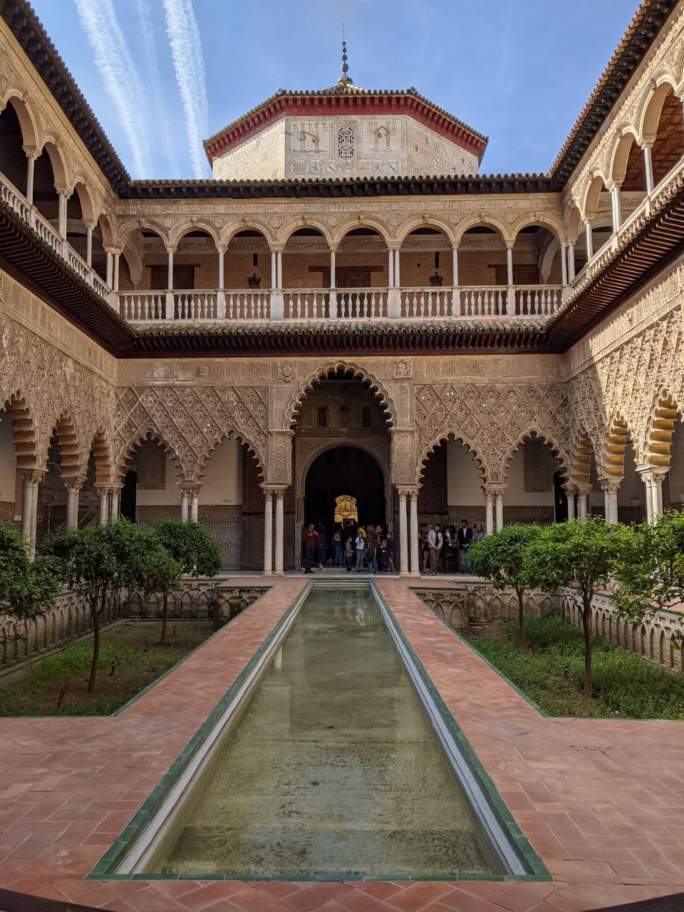 The Patio de las Doncellas in the Real Alcázar of Seville — a long rectangular reflecting pool framed by elaborately carved Mudéjar stucco arches and Renaissance upper galleries, with orange trees and visitors in the background
