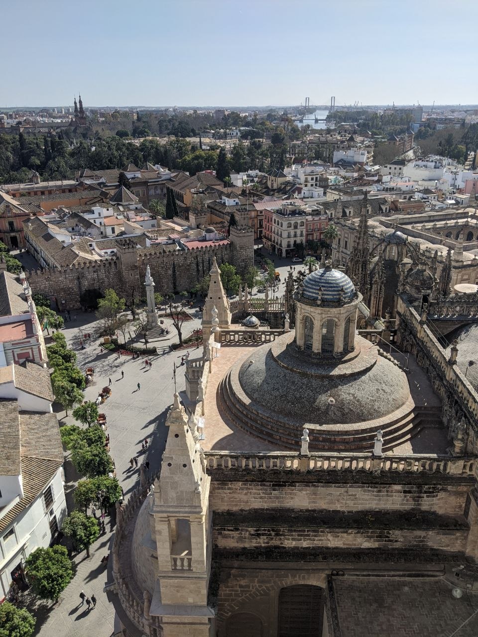Panoramic view from the top of the Giralda tower in Seville, looking out over the Gothic cathedral rooftop domes and pinnacles, the Alcázar walls, and the city stretching to the Guadalquivir River under a clear blue sky