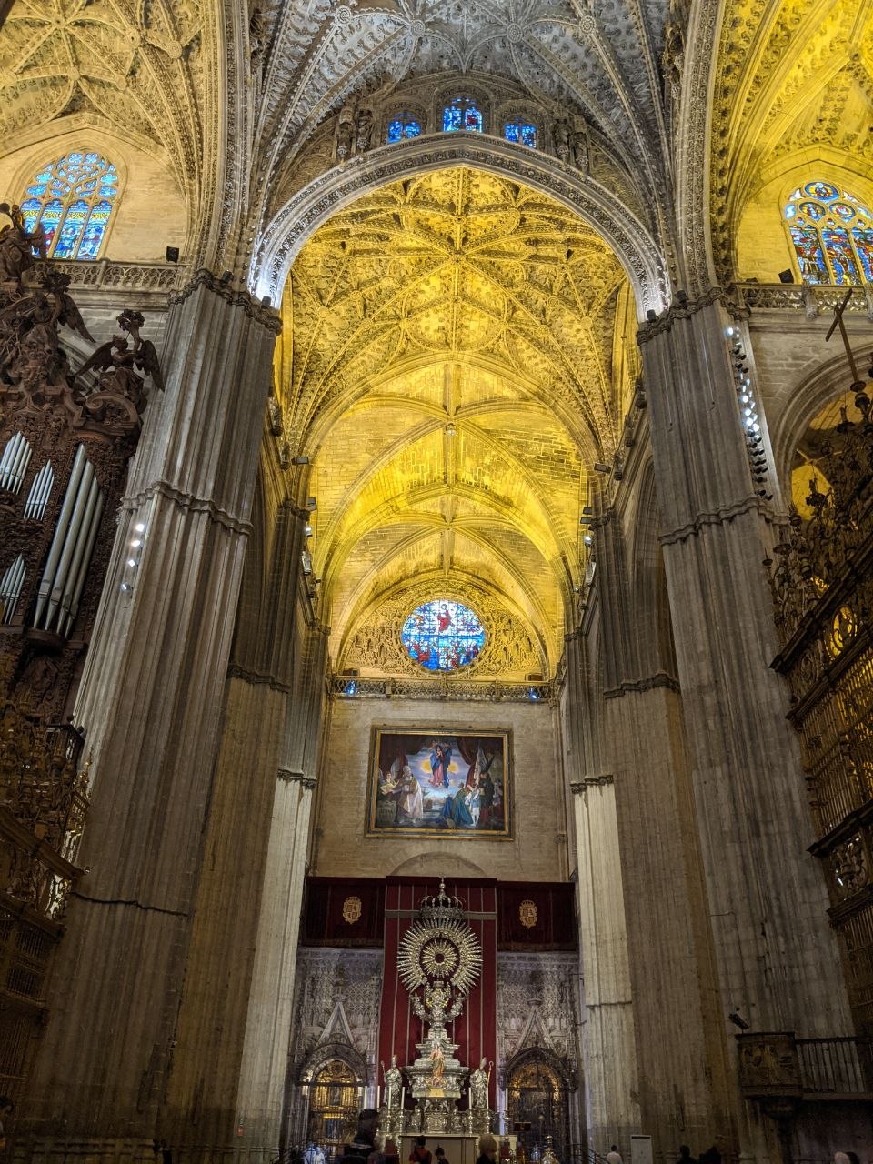 Interior of Seville Cathedral looking up at the illuminated golden ribbed vaulting of the main nave, with stained glass windows, pipe organ, and the ornate high altar visible below