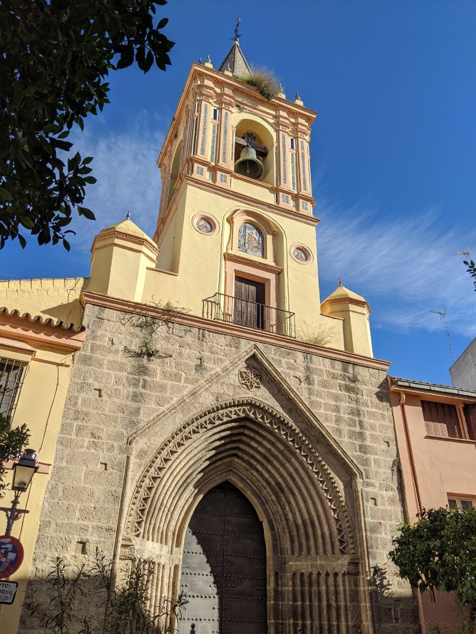 A Baroque church tower in Seville's historic centre rising above a medieval Gothic stone archway entrance, its cream plasterwork decorated with azulejo tile insets and a bell visible in the open belfry against a deep blue sky