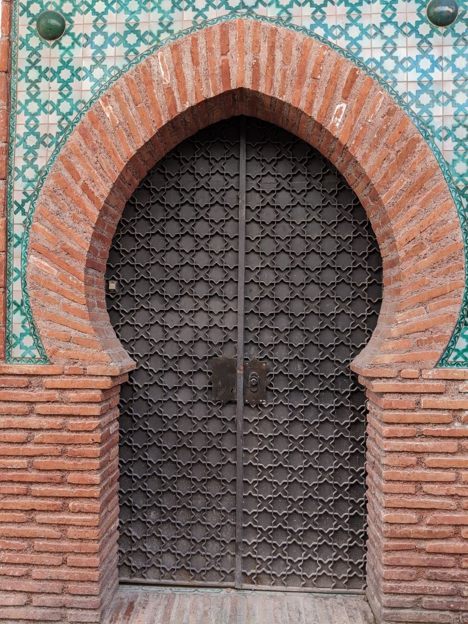 A Moorish horseshoe-arch doorway at the Grande Mosque of Granada, with intricate geometric iron grillwork gates set within a terracotta brick arch, the surrounding wall faced in green-and-white azulejo tilework
