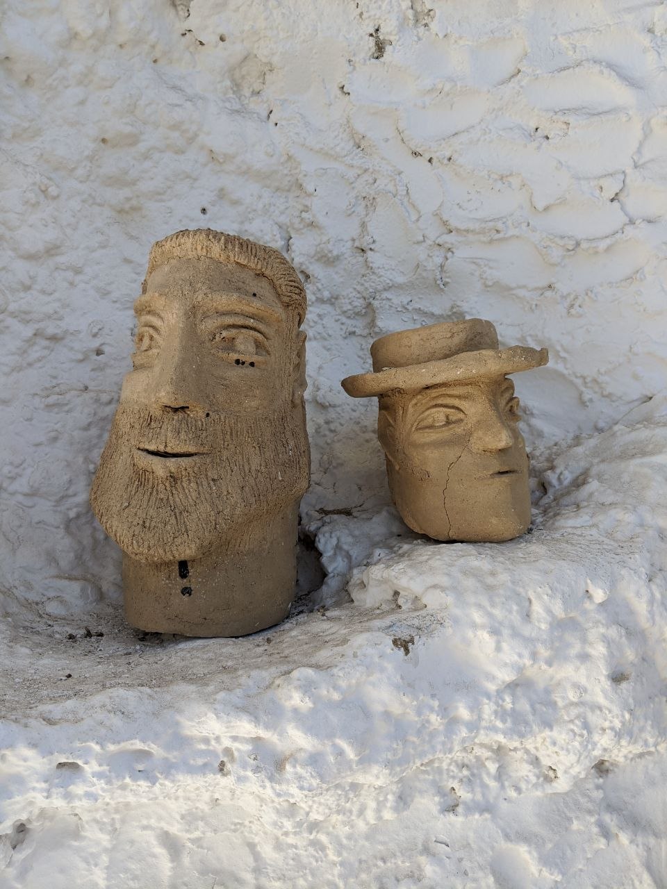Two hand-modelled terracotta clay head sculptures — one bearded, one wearing a wide-brimmed hat — resting on a ledge against the thick whitewashed wall of a Sacramonte cave house