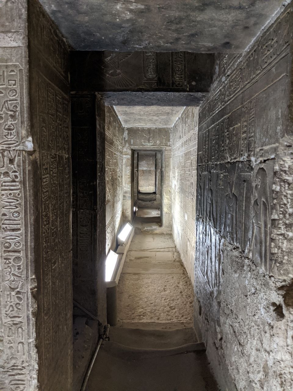 Narrow corridor of Abydos temple with carved hieroglyphs on walls and ceiling, lit by floor-level strip lights