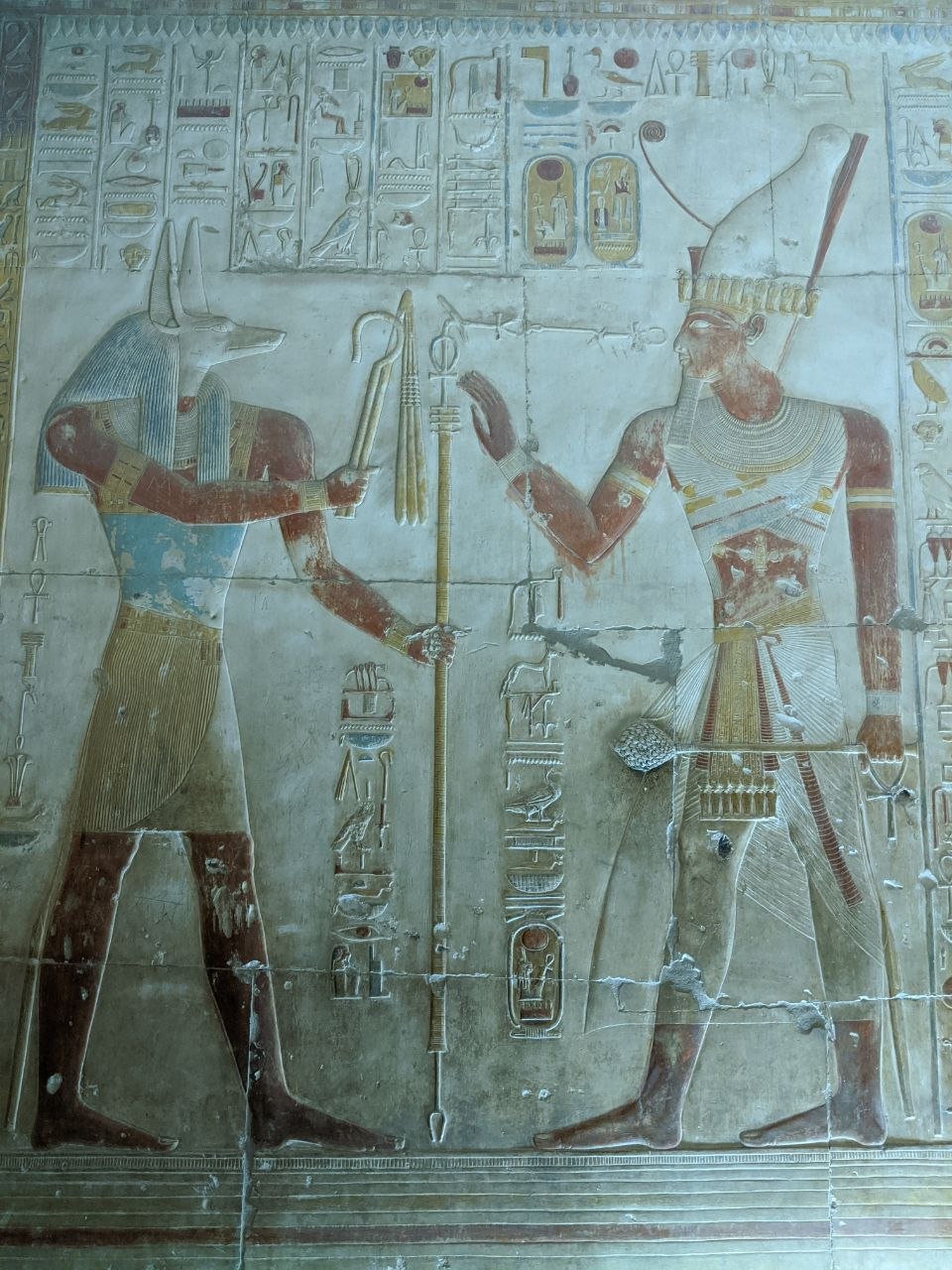 Vivid painted relief from Abydos showing Anubis facing a pharaoh wearing the white crown of Upper Egypt, both holding ceremonial staffs
