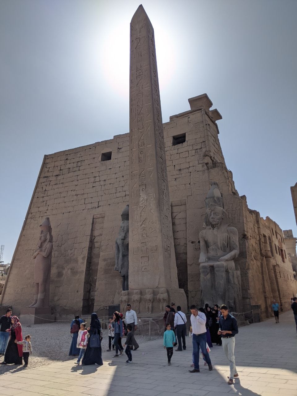 The obelisk of Ramesses II soaring against a hazy sky before the first pylon of Luxor Temple, flanked by colossal seated statues