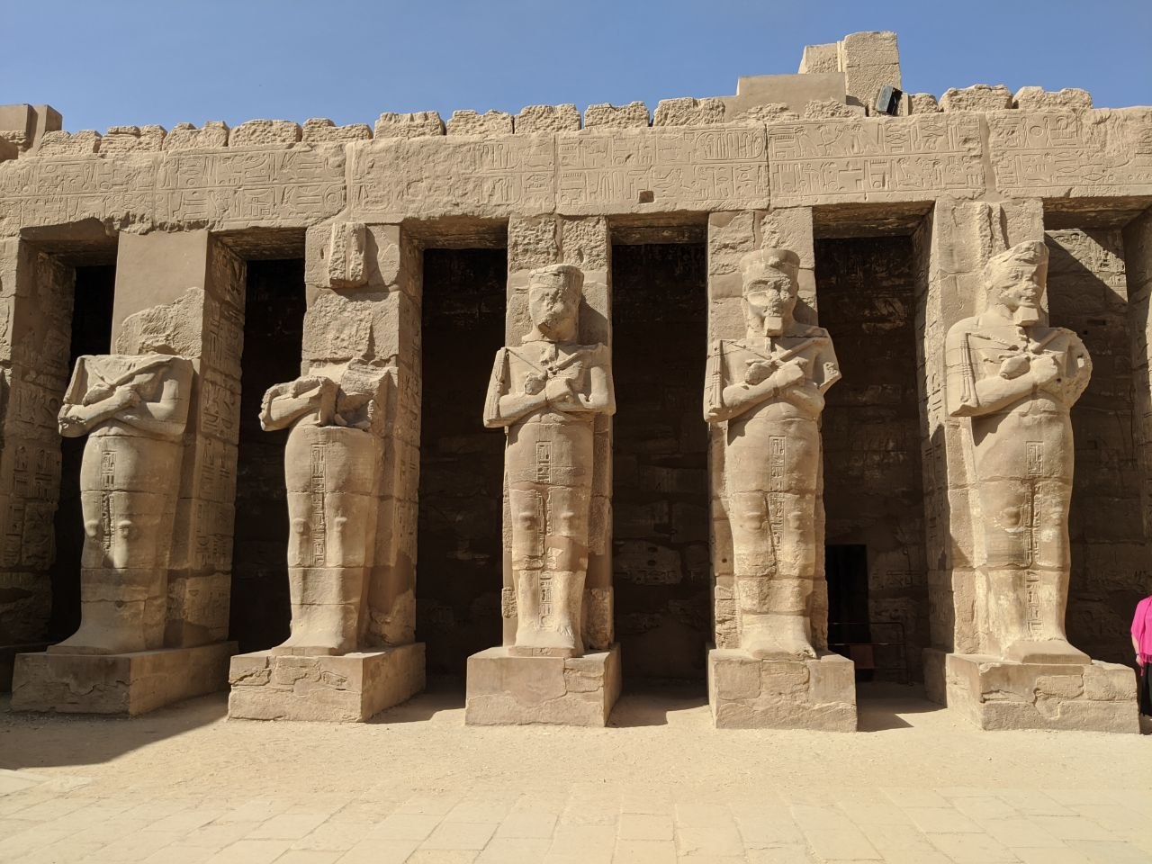 A row of Osiride statues of Ramesses II in mummiform, arms crossed holding crook and flail, standing before the walls of Karnak