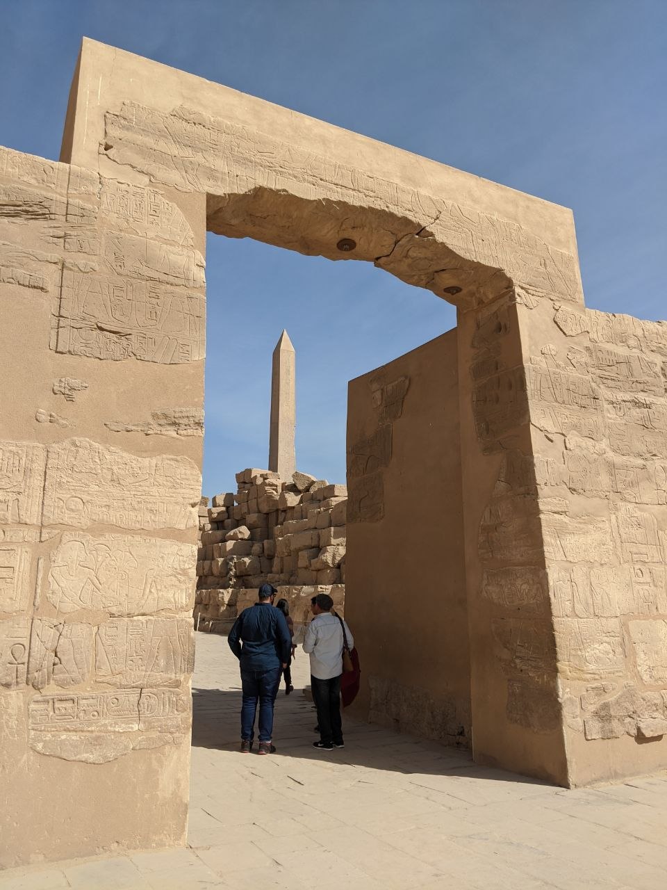 A massive gateway covered in carved hieroglyphs framing an obelisk in the distance, with visitors passing through