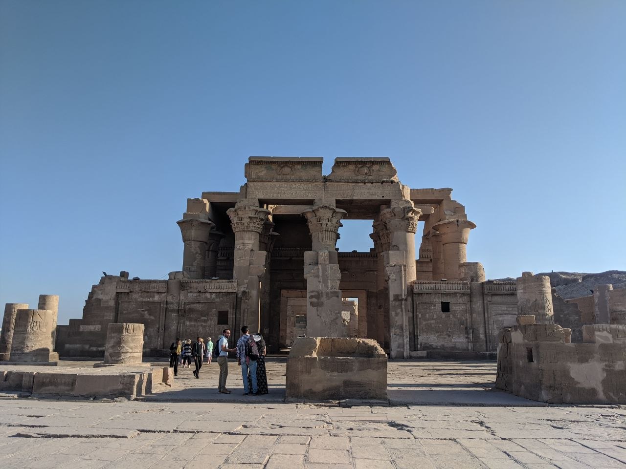 The double hypostyle hall of the Temple of Kom Ombo seen from the forecourt, its two parallel gateways framing columns with composite floral capitals under a clear blue sky, with visitors in the plaza