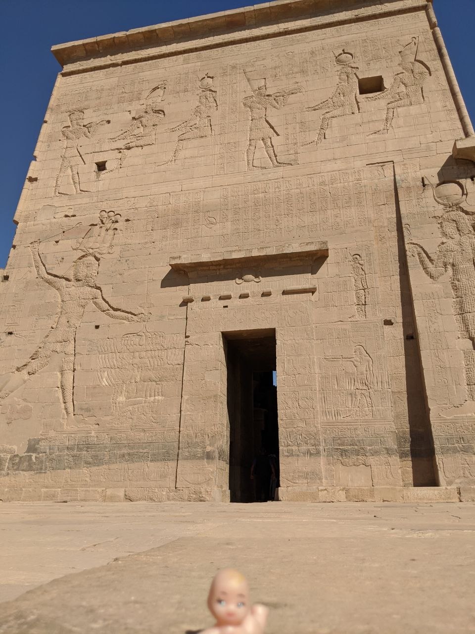 The main pylon of the Temple of Isis at Philae covered in large-scale carved relief showing pharaohs and deities, with a small doll figure in the foreground