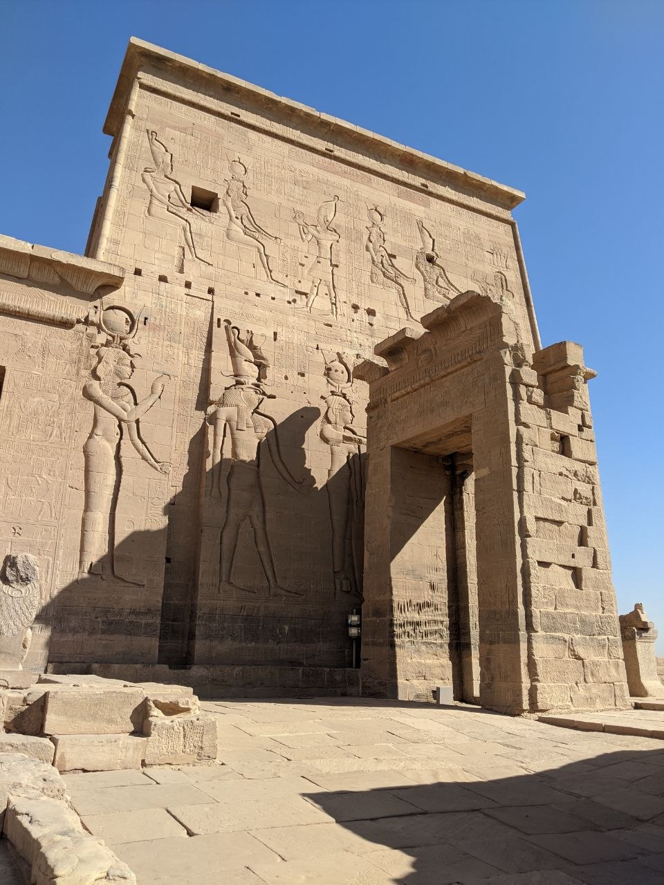 The inner pylon of Philae Temple with deeply carved relief figures of deities including Isis and Horus in sun, casting dramatic shadows across the sandstone surface