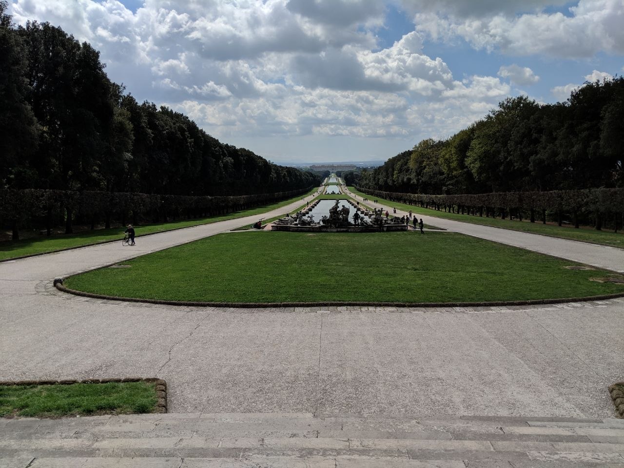 View along the main garden axis of the Royal Palace of Caserta, looking from the upper cascade toward the palace in the far distance, with tree-lined allées on either side, a central fountain group in the foreground, and visitors walking along the gravel path