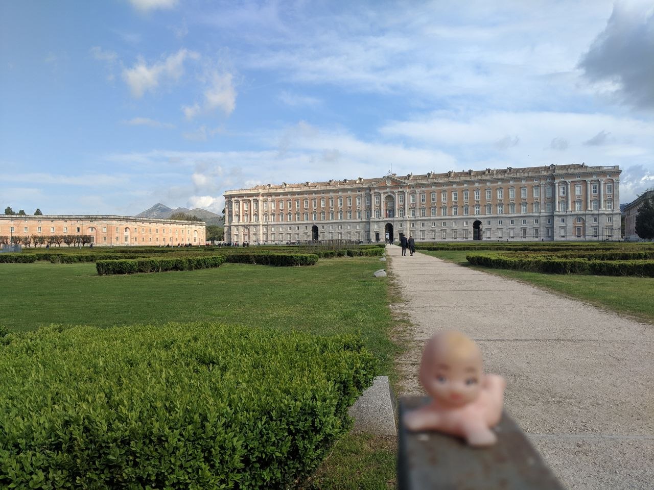 The long Baroque façade of the Royal Palace of Caserta seen from the formal gardens, with clipped box hedges in the foreground, a central gravel path leading to the entrance, and the Apennine hills visible behind