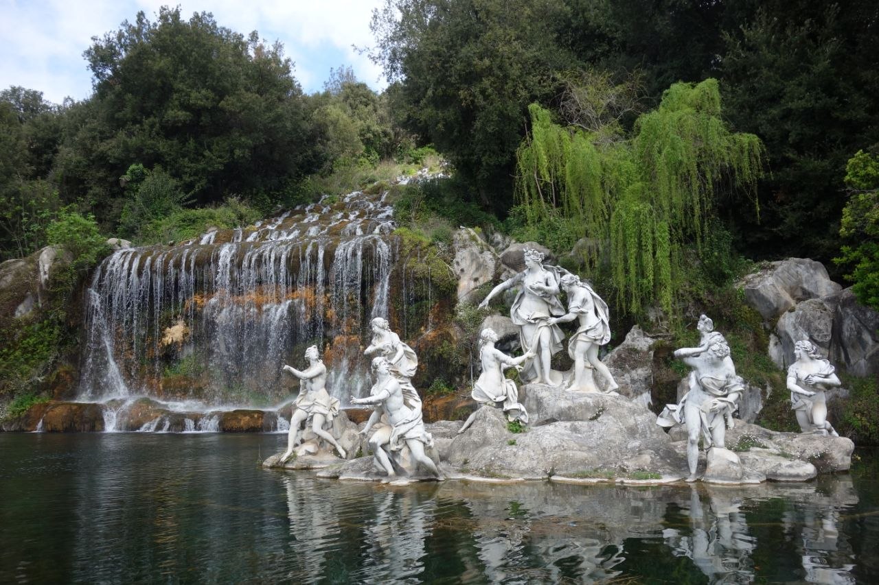 The Fountain of Diana and Actaeon at the Royal Palace of Caserta — a group of white marble mythological figures on a rocky island in a pool, with the grand cascade waterfall tumbling down the hillside behind them and willow trees framing the scene