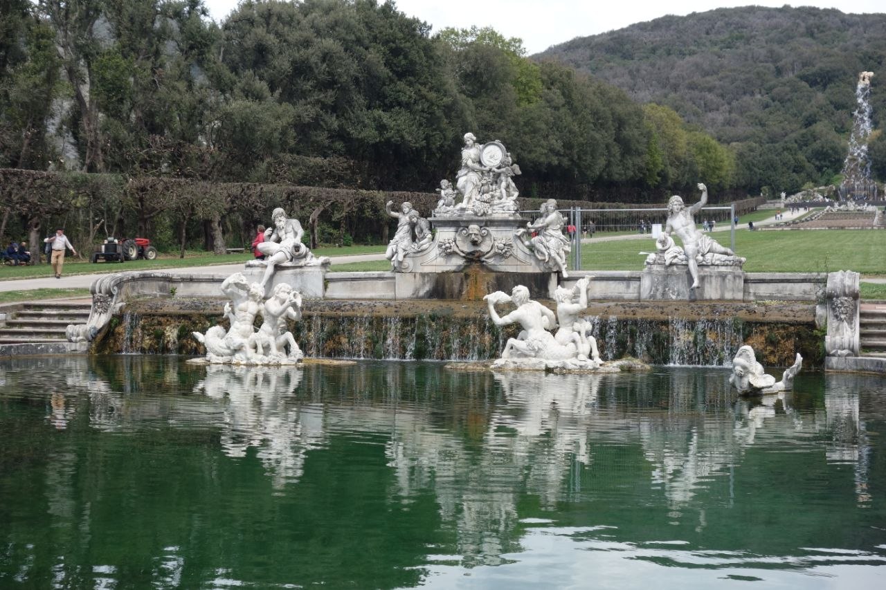 The Fountain of Ceres at the Royal Palace of Caserta — a tiered marble fountain with reclining river gods, tritons, and allegorical figures reflected in the dark green water of the basin, with the garden axis and hills visible beyond