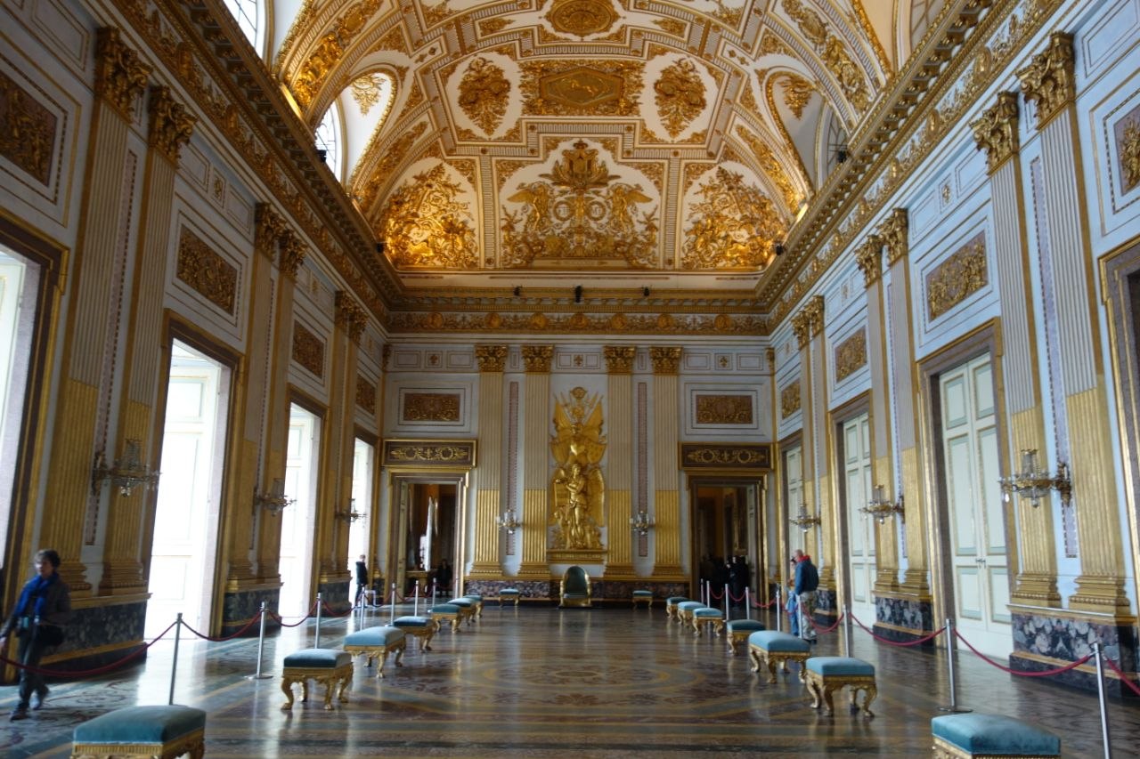 Interior of the Throne Room of the Royal Palace of Caserta — a long gilded hall with an elaborately decorated barrel-vaulted ceiling covered in gold stucco relief, flanked by pilasters and mirror-panelled doors, with a golden sculptural group above the throne at the far end and visitors along the sides