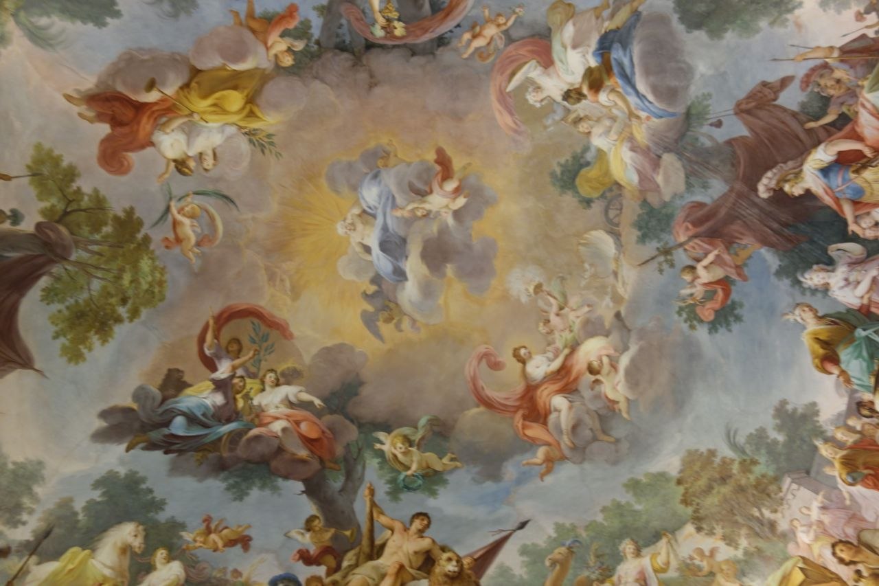 Baroque ceiling fresco inside the Royal Palace of Caserta depicting allegorical figures, gods, and putti swirling around a central luminous sky, with vivid colours and dramatic foreshortening across the entire painted vault