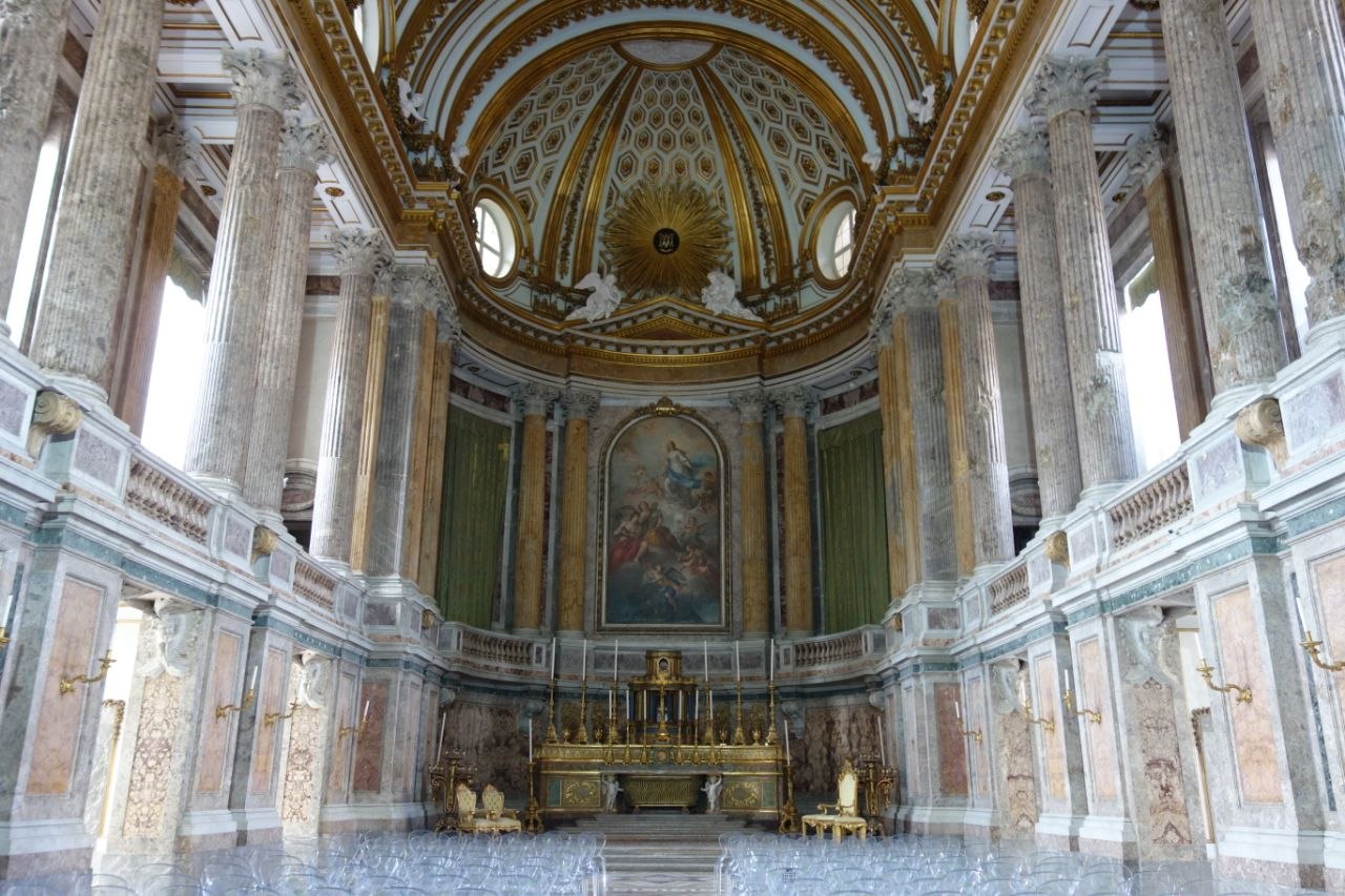 Interior of the palatine chapel of the Royal Palace of Caserta — a two-storey space with polychrome marble columns and walls in grey, green, and rose, a gilded coffered dome with a central sunburst, and the altarpiece of the Immaculate Conception above a gilt bronze altar