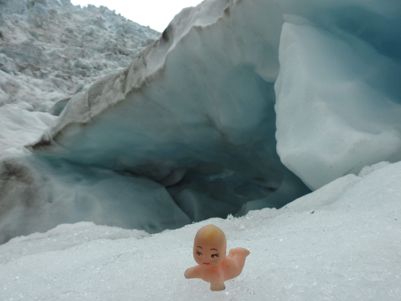 A small Kewpie doll figure placed on the white ice surface in front of a blue glacial ice cave entrance on Franz Josef Glacier, New Zealand, with the luminous turquoise interior of the crevasse visible behind