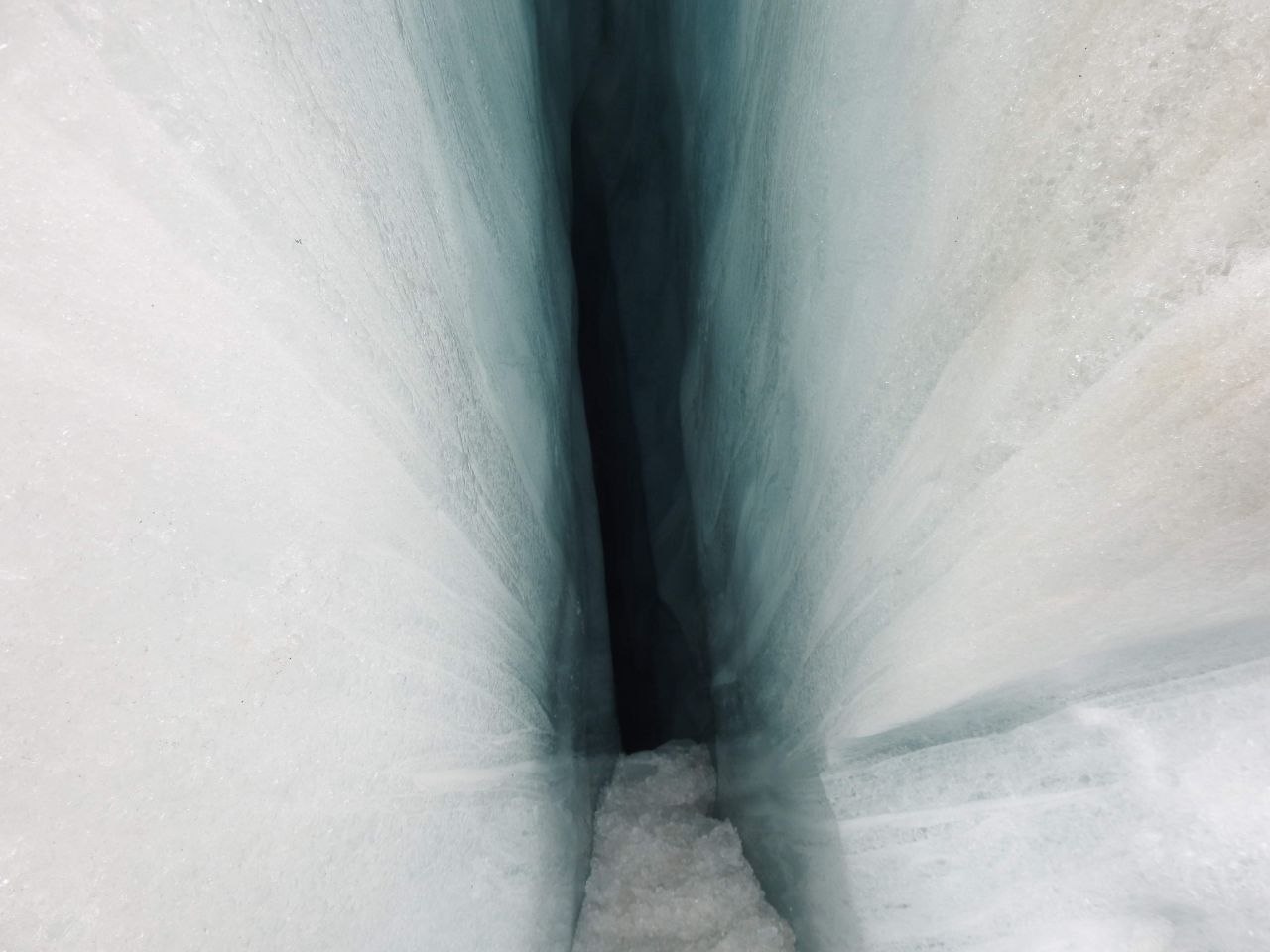 Close-up looking down into a deep glacial crevasse on Franz Josef Glacier, the walls of compressed ice glowing in shades of white and deep teal-blue, narrowing to darkness at the bottom