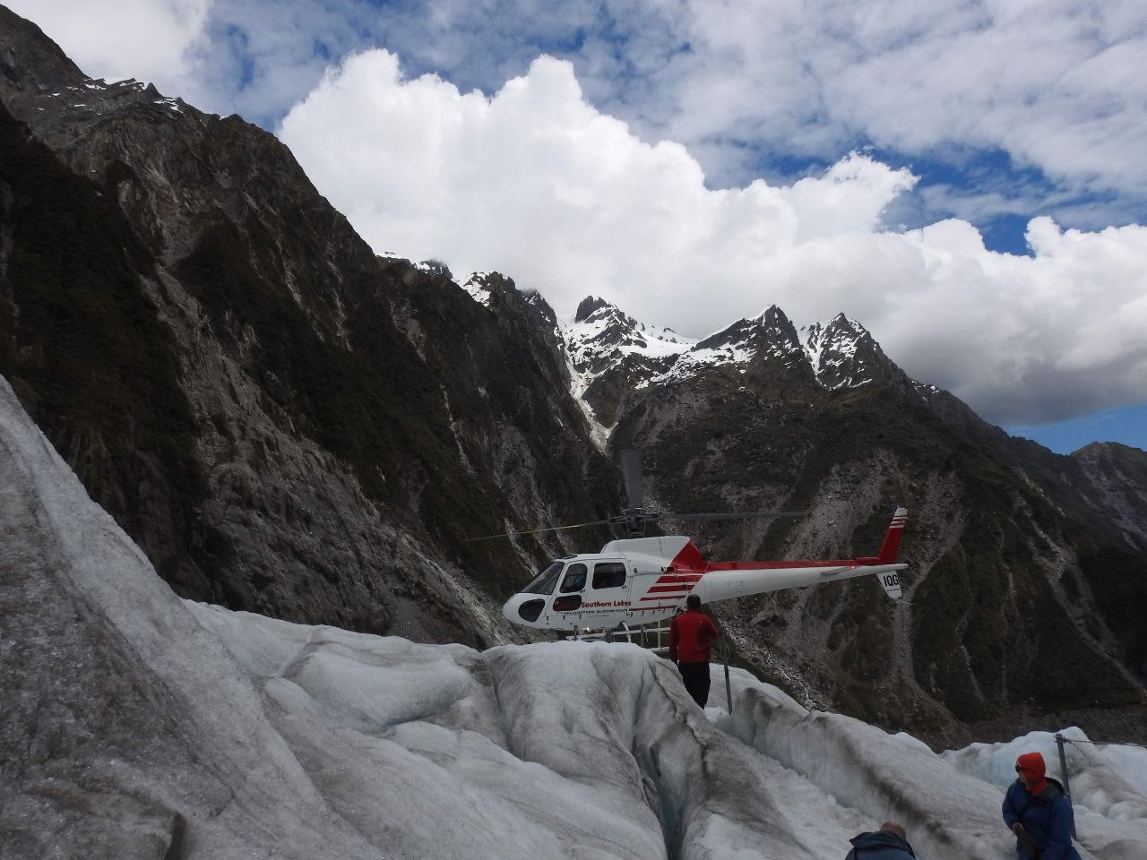 A Southern Lakes helicopter landed on the upper surface of Franz Josef Glacier between rocky valley walls, with snow-capped Southern Alps peaks rising into a partly cloudy sky behind and guides and passengers visible beside the aircraft