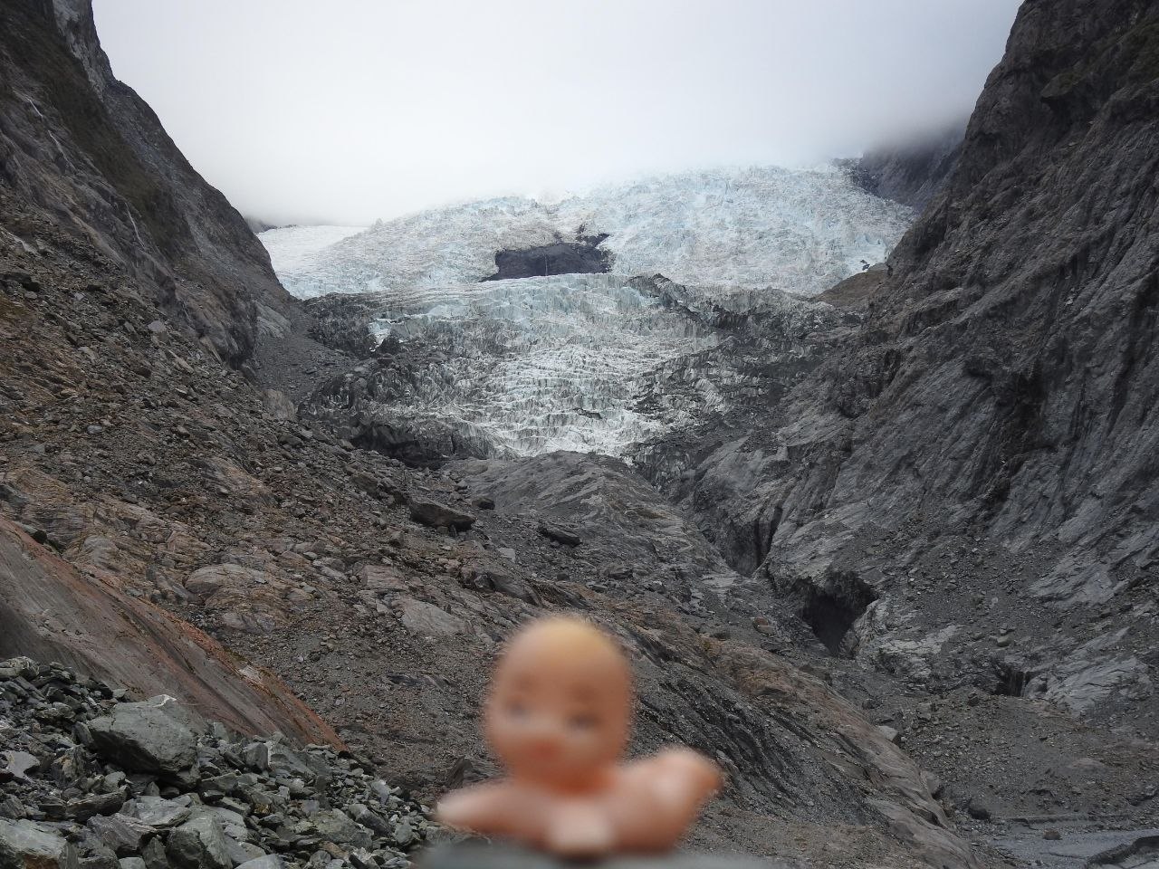 A small Kewpie doll figure held in the foreground facing the chaotic fractured face of Franz Josef Glacier, with its white and blue ice seracs filling the valley head between steep rocky walls under an overcast sky