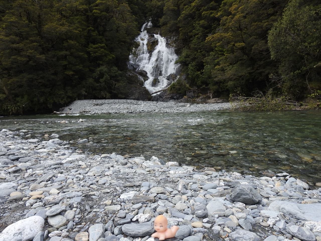 A Kewpie doll resting on the grey shingle riverbank beside the Blue Pools on the Makarora River, with a multi-tiered cascade waterfall tumbling over mossy rocks into the clear teal-green river behind, surrounded by beech forest