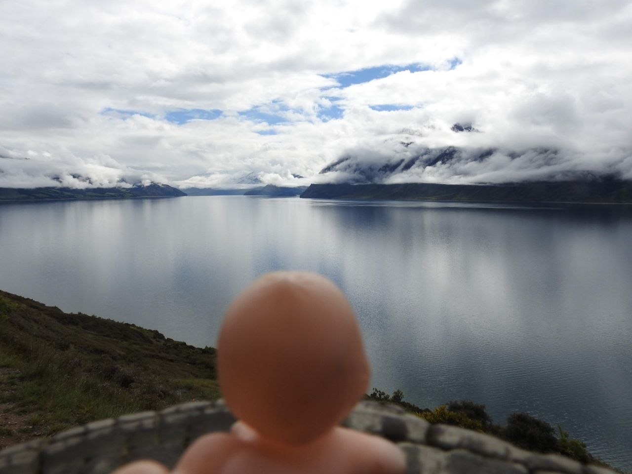 A Kewpie doll in the foreground on a tussock hillside above Lake Wānaka, its vast silver-grey surface perfectly still and reflecting cloud formations above, with the Southern Alps and Harris Mountains silhouetted in cloud on the far shore