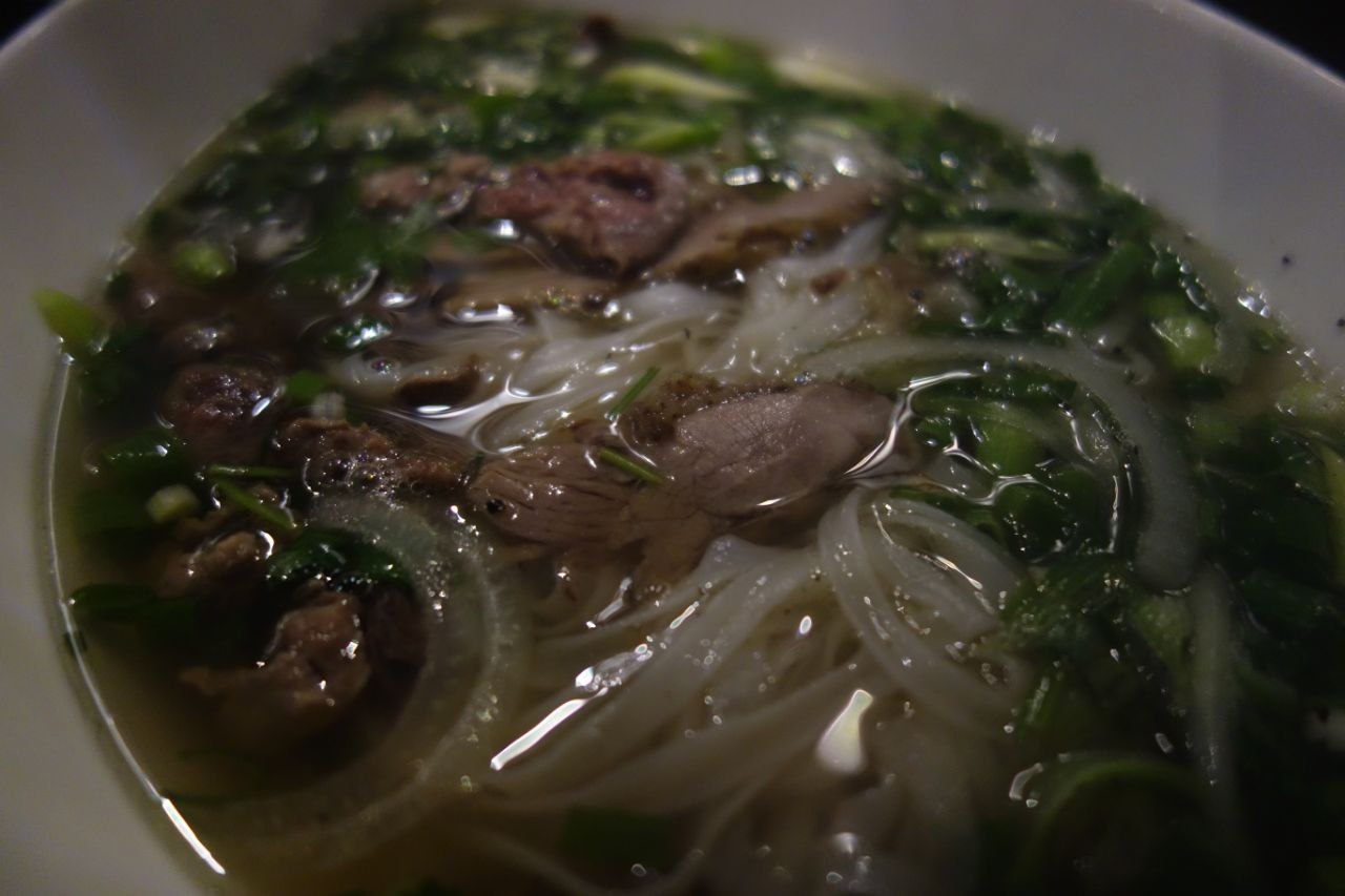 A large white bowl of Hanoi-style phở bò — the clear amber beef broth filled with flat rice noodles, slices of rare and well-done beef, white onion rings, and fresh herbs, shot from directly above in low light against a dark background