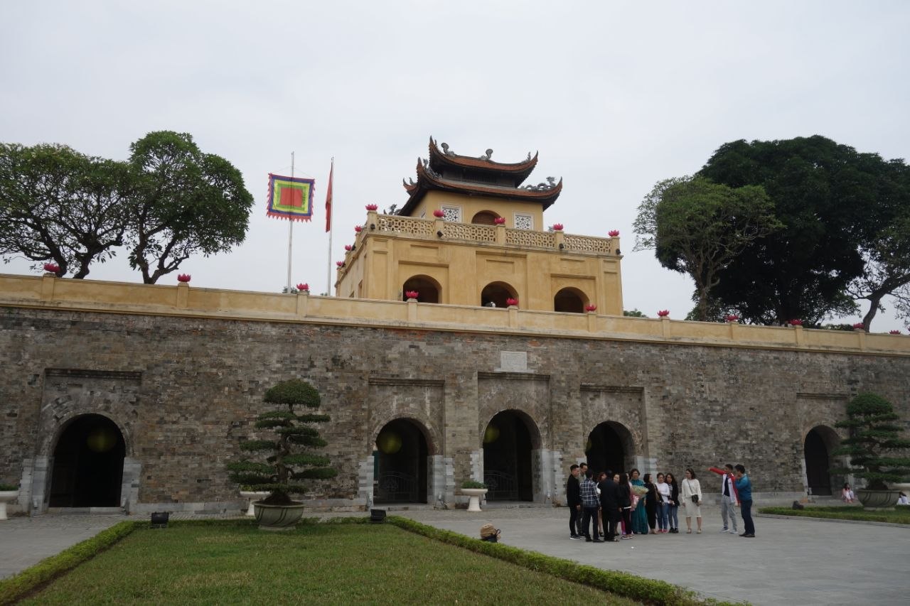 The Đoan Môn southern gate of the Imperial Citadel of Thăng Long in Hanoi — a wide masonry gatehouse of yellow-ochre stucco with five arched passageways through the lower level and a three-storey Vietnamese-style pavilion of upswept dark tiled roofs and red lotus finials rising above the battlemented wall, flanked by bonsai pines, with a group of visitors in the foreground and the Vietnamese and Communist Party flags flying against an overcast grey sky