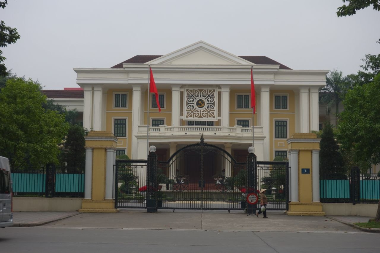 A French colonial-era government building in the Ba Đình district of Hanoi — a symmetrical two-storey yellow and white neoclassical facade with a central pediment, Ionic pilasters, and a large decorative communist emblem (hammer and sickle within a geometric lattice frame) mounted on the upper floor between two red Vietnamese flags, a wrought-iron gate in front, a woman in a conical hat walking past on the empty street below under an overcast sky