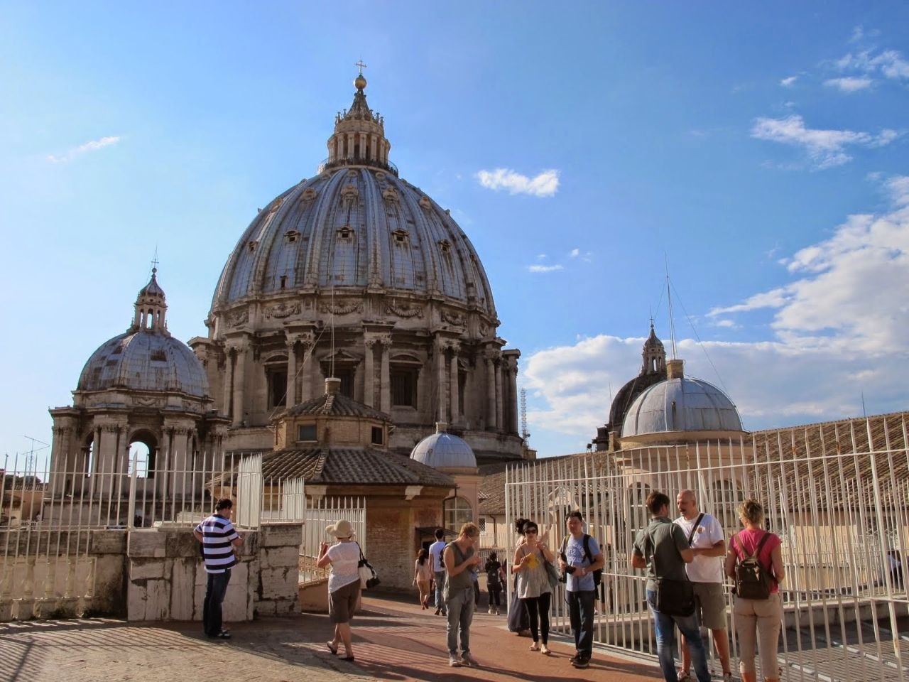 The dome of St. Peter's Basilica seen from the basilica rooftop, with visitors on the terrace and two smaller domes flanking it against a blue sky