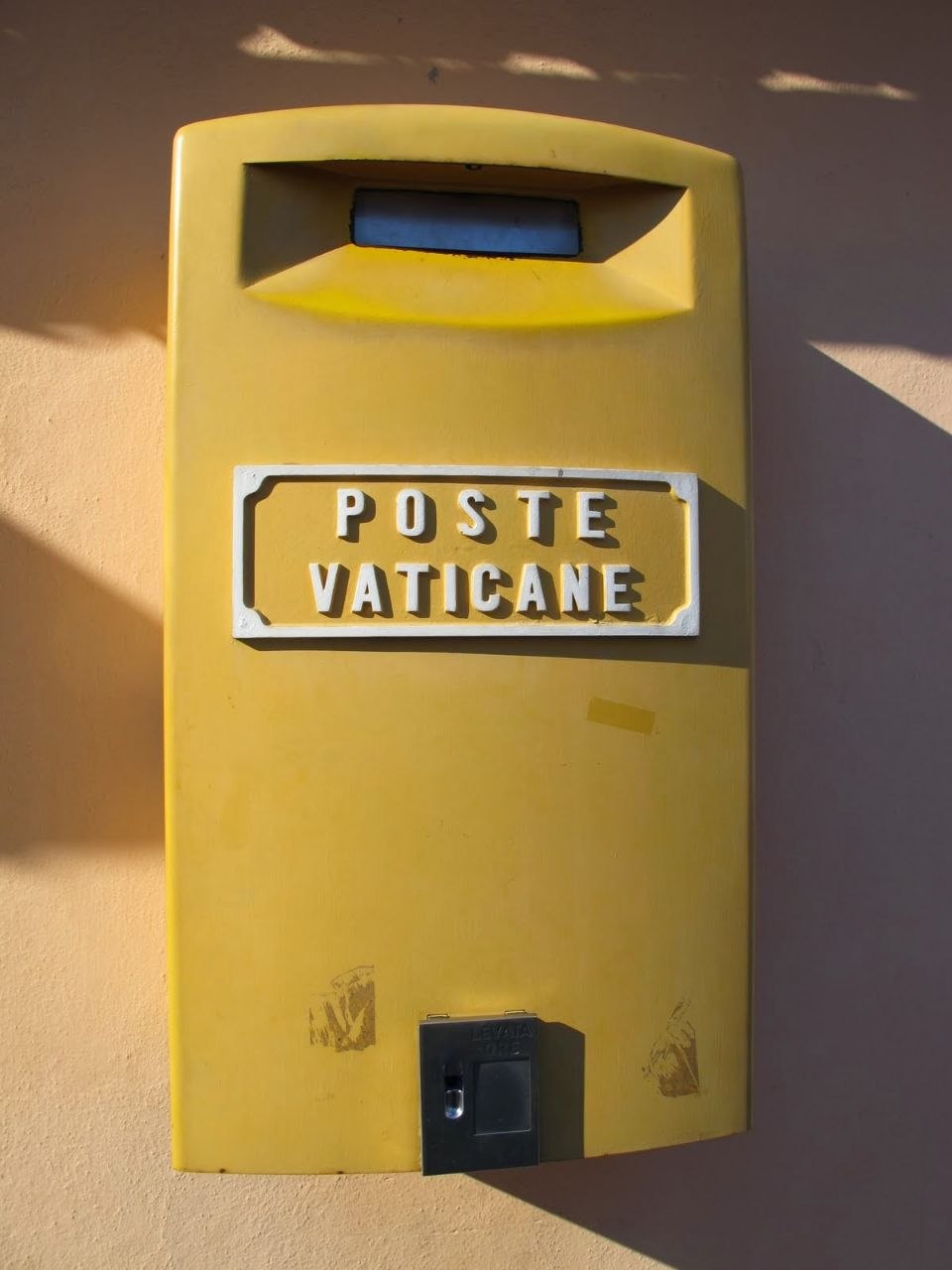 A yellow Poste Vaticane postbox mounted on a pale ochre wall in Vatican City, casting a strong shadow in bright sunlight