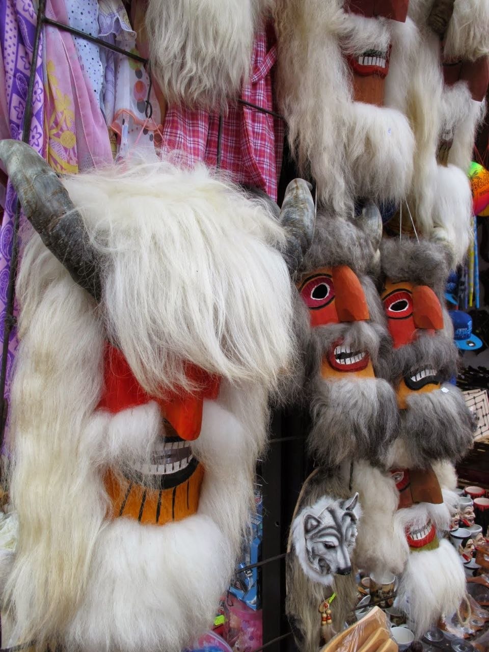 Traditional Romanian carnival masks displayed at an outdoor market stall near Bran — large carved wooden faces with exaggerated orange beaks and grinning teeth, covered in thick white and grey animal fur with curling ram horns, hung alongside colourful folk costumes and smaller ceramic souvenirs