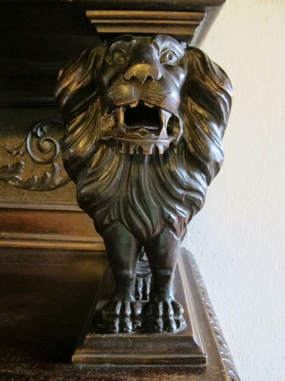 A dark carved wooden lion corbel inside Bran Castle — a highly detailed roaring lion's head with an open mouth and flowing mane, supporting a heavy dark timber mantelpiece, with ornate foliate scrollwork visible to the left, against a plain cream plaster wall